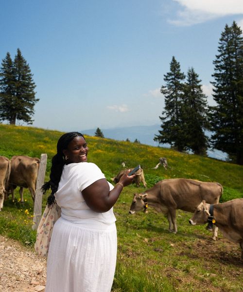 A person wearing a white outfit smiles and holds a phone while standing in a grassy field with cows, under a bright blue sky with trees in the background.