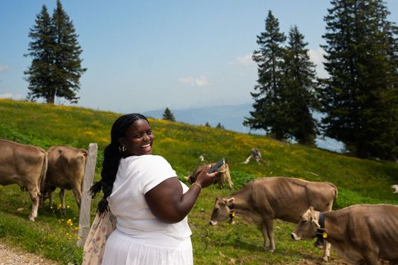 A person wearing a white outfit smiles and holds a phone while standing in a grassy field with cows, under a bright blue sky with trees in the background.