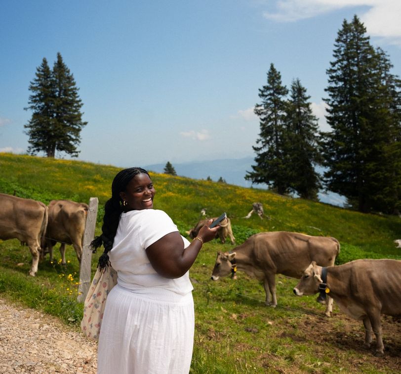 A person wearing a white outfit smiles and holds a phone while standing in a grassy field with cows, under a bright blue sky with trees in the background.