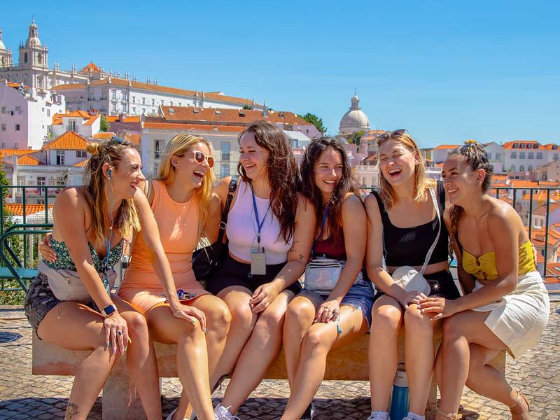 Six women sitting on a bench, laughing and enjoying a sunny day with a scenic cityscape in the background.