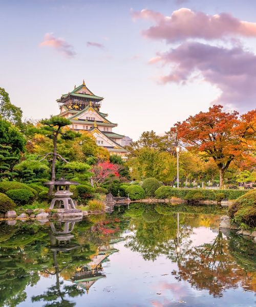 A traditional Japanese castle surrounded by lush gardens with colorful autumn trees, reflected in a serene pond under a pastel sky.