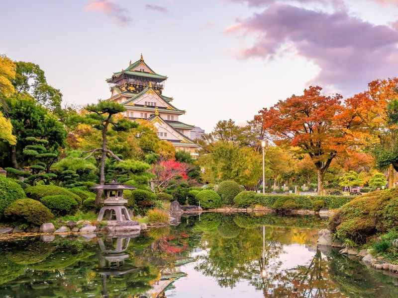 A traditional Japanese castle surrounded by lush gardens with colorful autumn trees, reflected in a serene pond under a pastel sky.