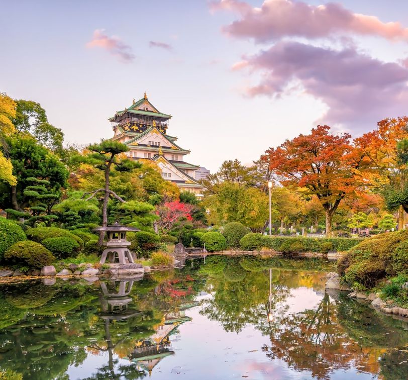 A traditional Japanese castle surrounded by lush gardens with colorful autumn trees, reflected in a serene pond under a pastel sky.