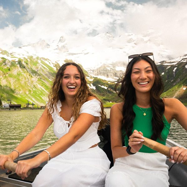 Two women smiling while rowing a boat on a mountain lake, with green hills and snowy peaks in the background.