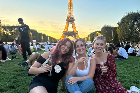 Three women sit on the grass holding drinks, with the Eiffel Tower illuminated in the background at sunset.