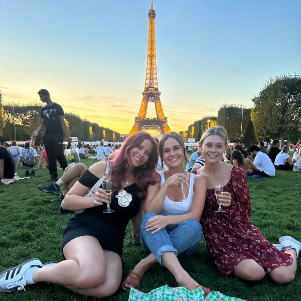 Three women sit on the grass holding drinks, with the Eiffel Tower illuminated in the background at sunset.