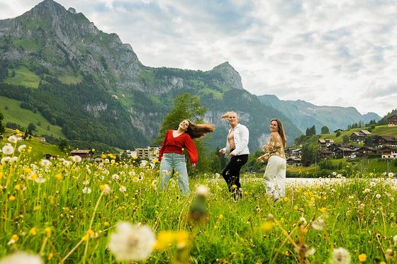 Three people dance joyfully in a lush meadow with dandelions, backed by majestic mountains and a quaint village under a cloudy sky.