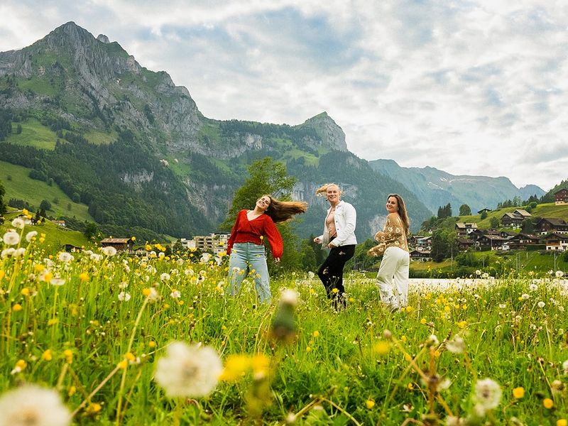 Three people dance joyfully in a lush meadow filled with wildflowers, with mountainous scenery and a cloudy sky in the background.