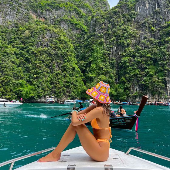 A woman in a bikini and colorful hat sits on a boat's bow, surrounded by turquoise water and lush, rocky cliffs.
