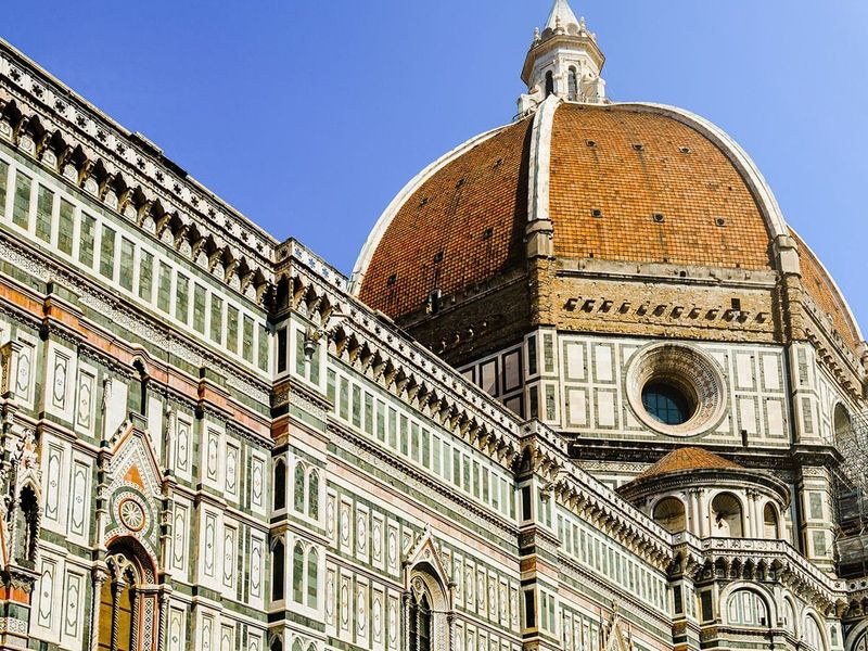 The image shows the Florence Cathedral with its iconic red-tiled dome, intricate green and white marble facade, and a clear blue sky in the background.