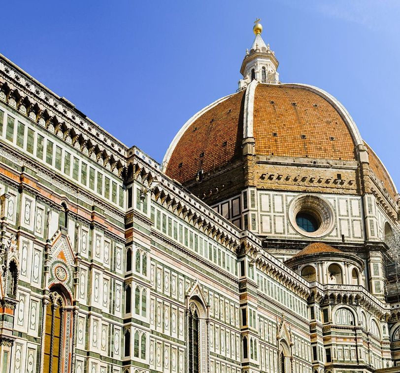The image shows the Florence Cathedral with its iconic red-tiled dome, intricate green and white marble facade, and a clear blue sky in the background.