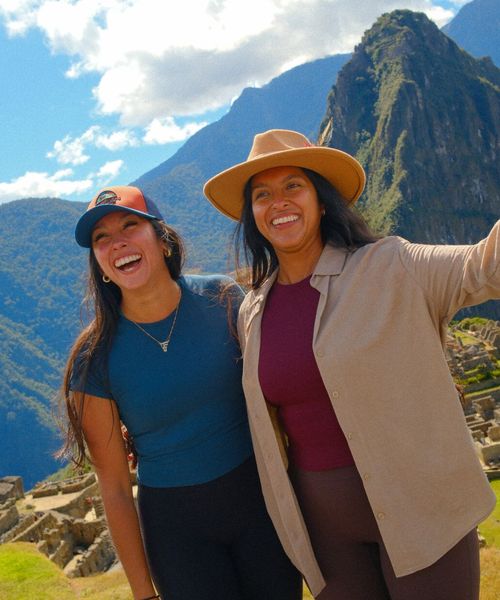 Two women smiling and posing in front of Machu Picchu, with the iconic green mountain and ruins visible under a bright, partly cloudy sky.