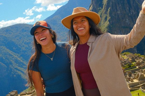 Two women smiling and posing in front of Machu Picchu, with the iconic green mountain and ruins visible under a bright, partly cloudy sky.