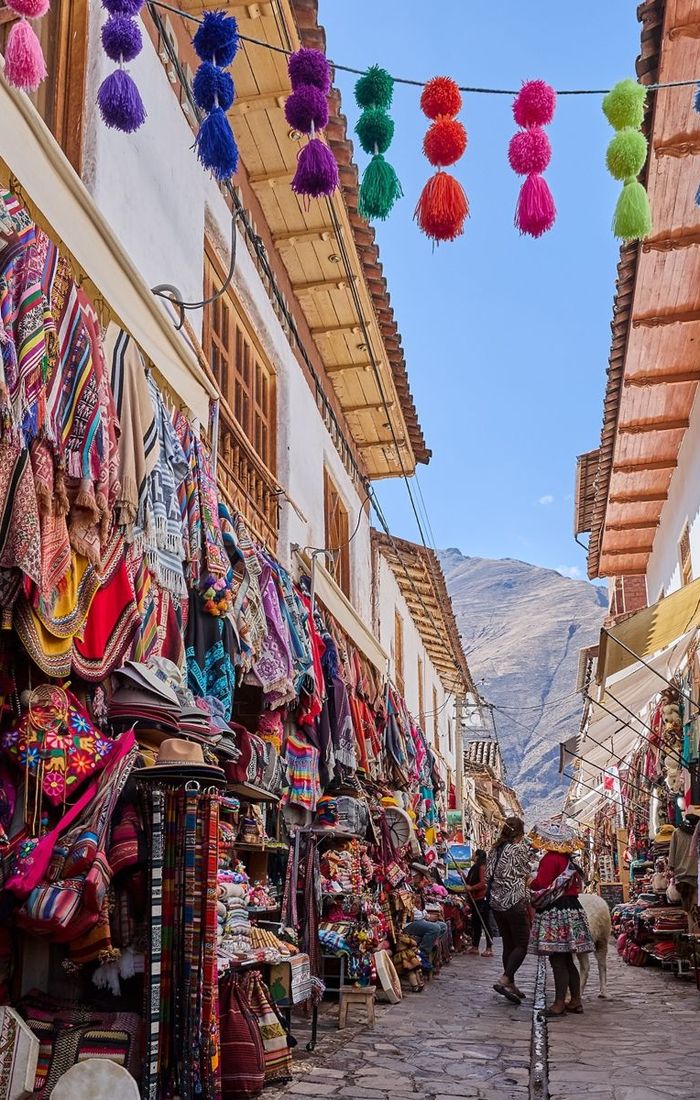 A vibrant street market with colorful textiles and garments displayed, traditional decorations hanging overhead, and people walking along the cobblestone path.