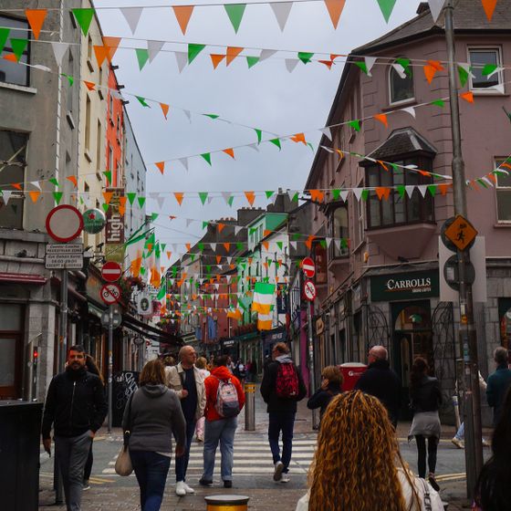 Busy street decorated with green, white, and orange flags, bustling with people, surrounded by colorful buildings and overcast sky.