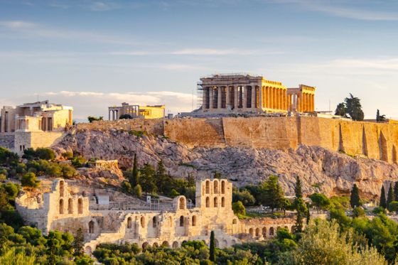 The Parthenon atop the Acropolis in Athens, Greece, bathed in golden sunlight, surrounded by lush greenery and ancient ruins.