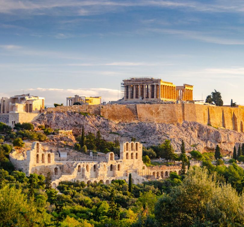 The Parthenon atop the Acropolis in Athens, Greece, bathed in golden sunlight, surrounded by lush greenery and ancient ruins.