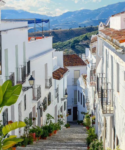 Narrow, picturesque street in a whitewashed village with potted plants and mountain views in the background under a blue sky.