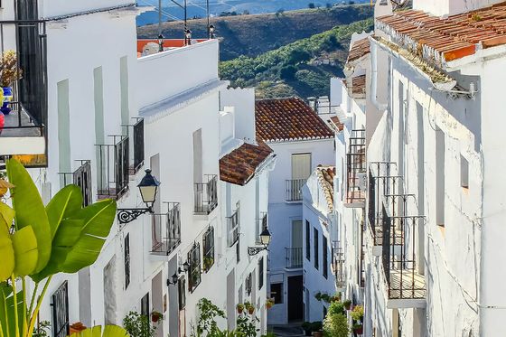 Narrow, picturesque street in a whitewashed village with potted plants and mountain views in the background under a blue sky.