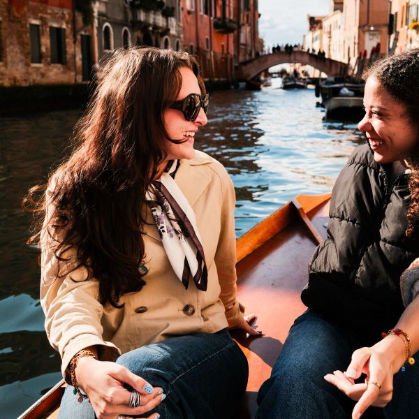 Two women smiling and talking while sitting in a boat on a canal, with historic buildings and a bridge in the background.