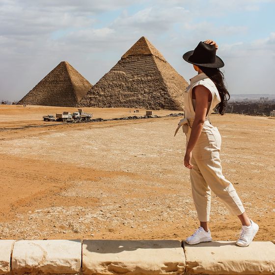 Woman in beige outfit and hat stands facing away, looking at the Pyramids of Giza under a cloudy sky.