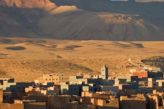 A desert village with clay buildings and a mosque tower, surrounded by palm trees, set against a backdrop of rugged mountains under a blue sky.