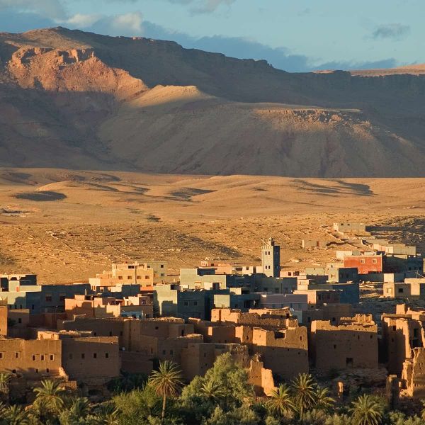 A desert village with clay buildings and a mosque tower, surrounded by palm trees, set against a backdrop of rugged mountains under a blue sky.
