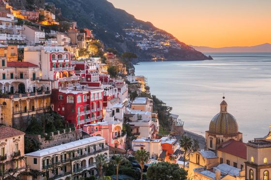 A coastal village with vibrant cliffside houses overlooking the sea at sunset, featuring a dome-roofed church in the foreground.