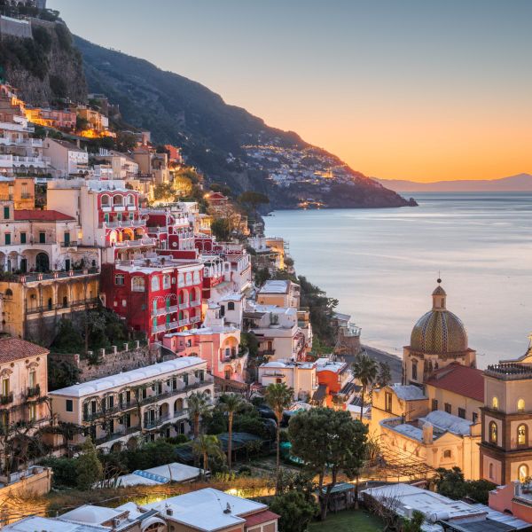 A coastal village with vibrant cliffside houses overlooking the sea at sunset, featuring a dome-roofed church in the foreground.