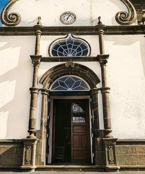 Ornate church entrance with arched wooden door, decorative stone columns, and circular window under a clock on a sunlit facade.