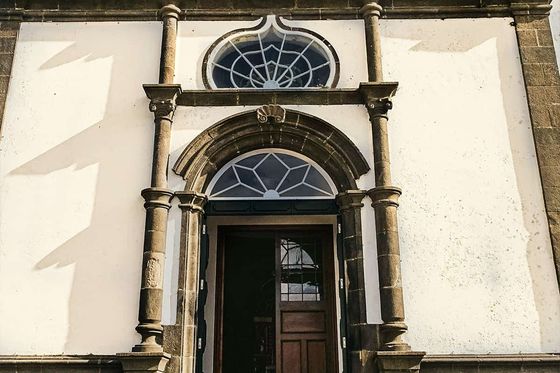 Ornate church entrance with arched wooden door, decorative stone columns, and circular window under a clock on a sunlit facade.