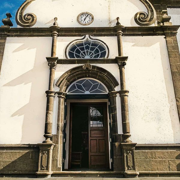 Ornate church entrance with arched wooden door, decorative stone columns, and circular window under a clock on a sunlit facade.