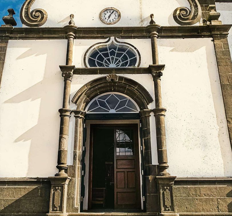 Ornate church entrance with arched wooden door, decorative stone columns, and circular window under a clock on a sunlit facade.