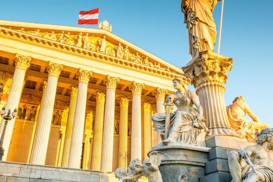 A grand neoclassical building with a statue in front, featuring ornate sculptures and columns, lit by golden sunlight, with a flag on top.