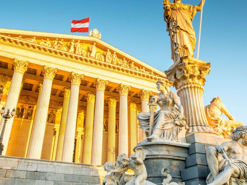 A grand neoclassical building with a statue in front, featuring ornate sculptures and columns, lit by golden sunlight, with a flag on top.