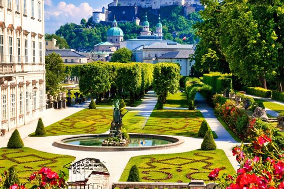 Beautiful garden with manicured hedges and a statue, leading to a historic fortress in the background, under a clear blue sky.