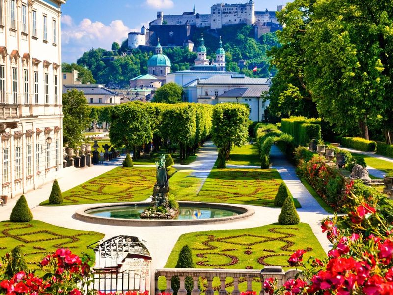Beautiful garden with manicured hedges and a statue, leading to a historic fortress in the background, under a clear blue sky.