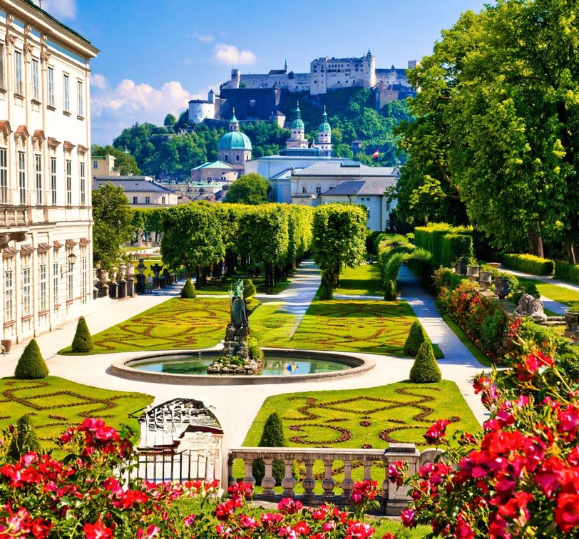 Beautiful garden with manicured hedges and a statue, leading to a historic fortress in the background, under a clear blue sky.