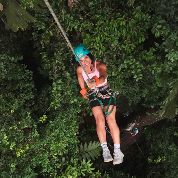 A person wearing a helmet and safety harness is ziplining through dense, green forest foliage, smiling at the camera.