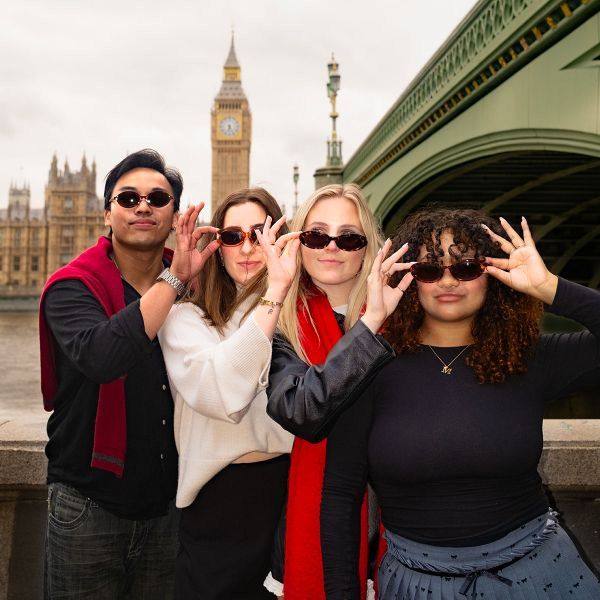 Four people in sunglasses pose together with the Big Ben and Westminster Bridge in the background on a cloudy day.