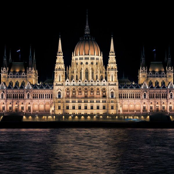 Illuminated Gothic-style building with towers and a dome, reflecting on a river at night.