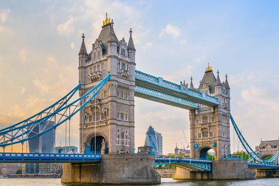Tower Bridge in London spans the River Thames, with its iconic twin towers and blue suspension cables under a partly cloudy sky.