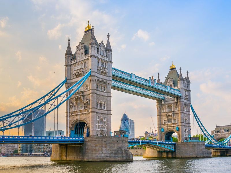 Tower Bridge in London spans the River Thames, with its iconic twin towers and blue suspension cables under a partly cloudy sky.