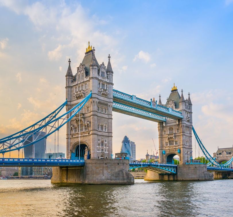 Tower Bridge in London spans the River Thames, with its iconic twin towers and blue suspension cables under a partly cloudy sky.
