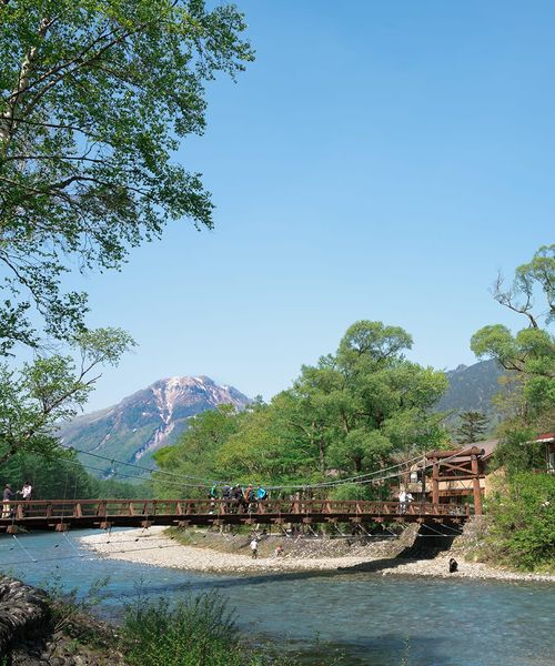 A scenic view of a wooden bridge over a clear river, surrounded by lush greenery and mountains under a bright blue sky.