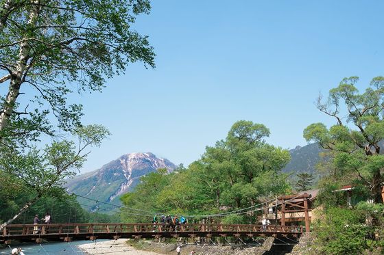 A scenic view of a wooden bridge over a clear river, surrounded by lush greenery and mountains under a bright blue sky.