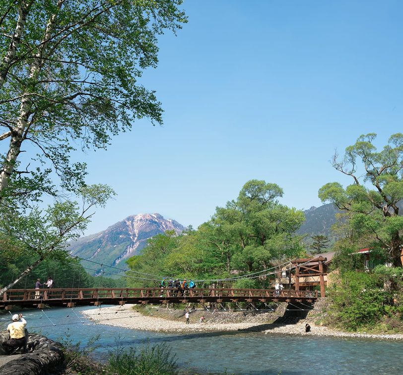 A scenic view of a wooden bridge over a clear river, surrounded by lush greenery and mountains under a bright blue sky.