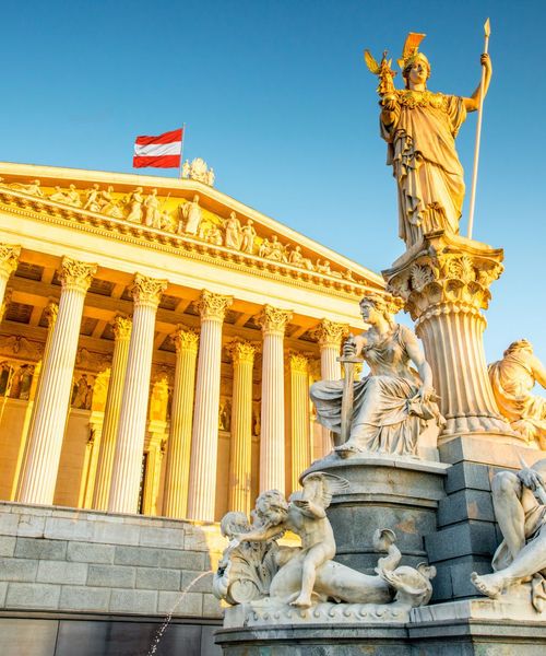 Historic building with Corinthian columns and statue, adorned with detailed sculptures, under a clear blue sky and a flag waving on top.