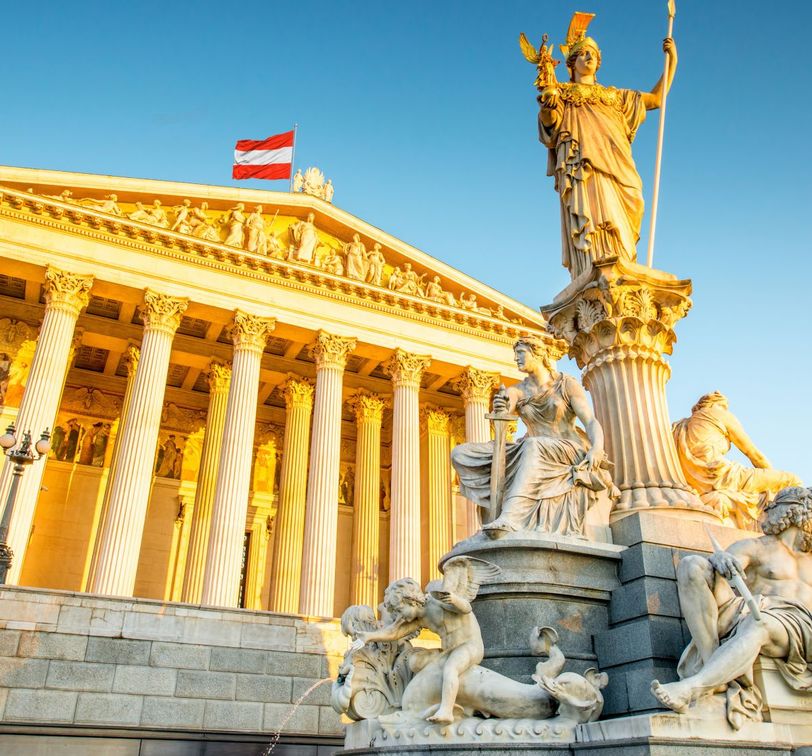 Historic building with Corinthian columns and statue, adorned with detailed sculptures, under a clear blue sky and a flag waving on top.