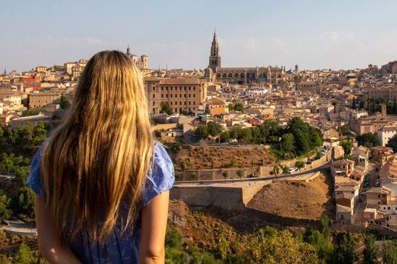 A woman with long blonde hair stands overlooking a historic cityscape under a clear blue sky.
